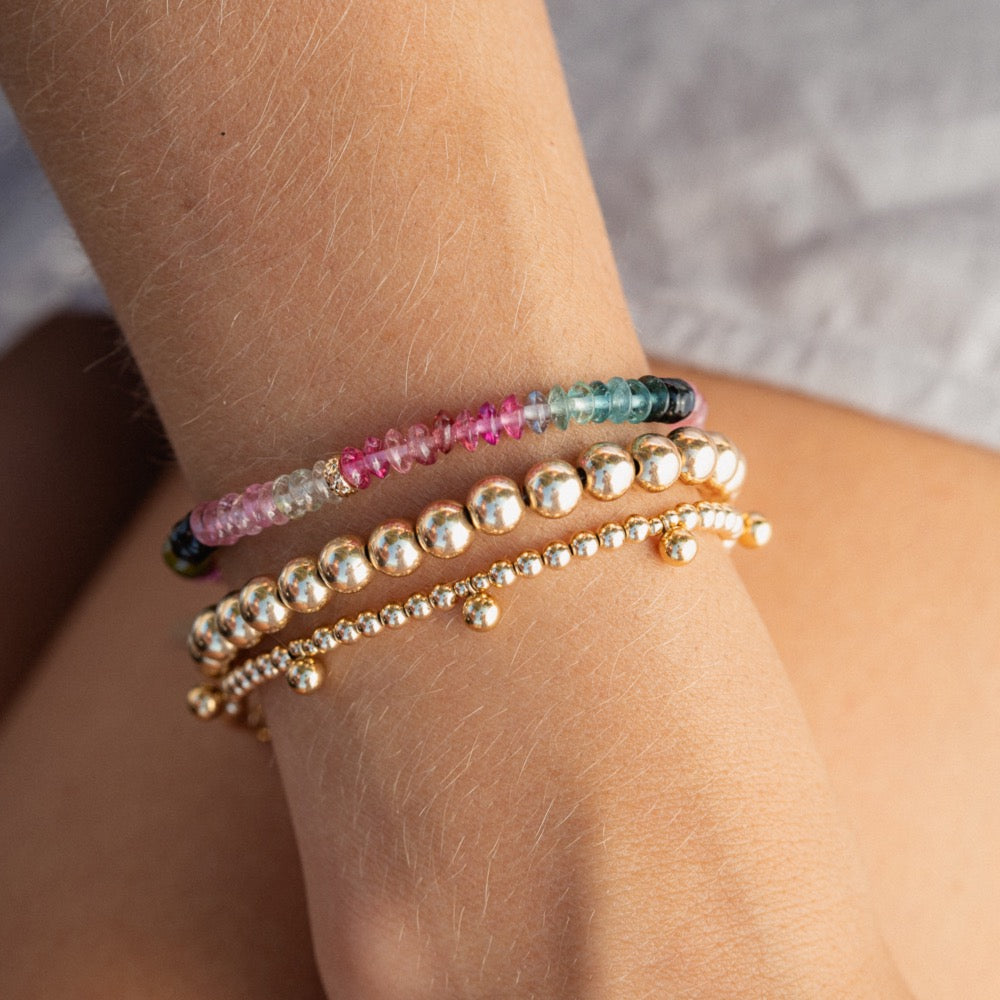 A close-up view of a person's wrist wearing multiple bracelets, including a beaded bracelet with a row of gemstones and a gold-toned bracelet with small round beads.